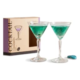 Two clear glass cocktail glasses with green liquid on a wooden table next to a plant and product box, with sofa in backgroun… - view 2
