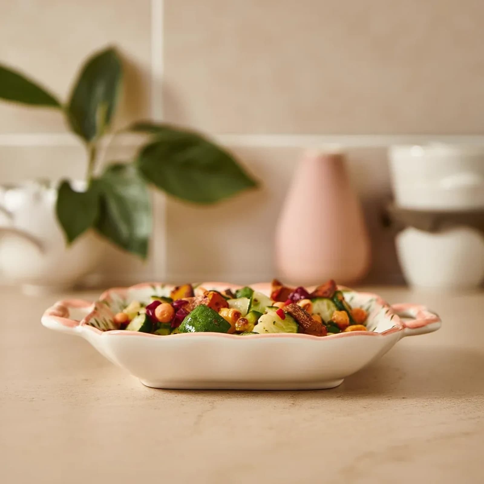 White porcelain serving dish with pink wavy edges and floral pattern, filled with salad, on a kitchen counter.