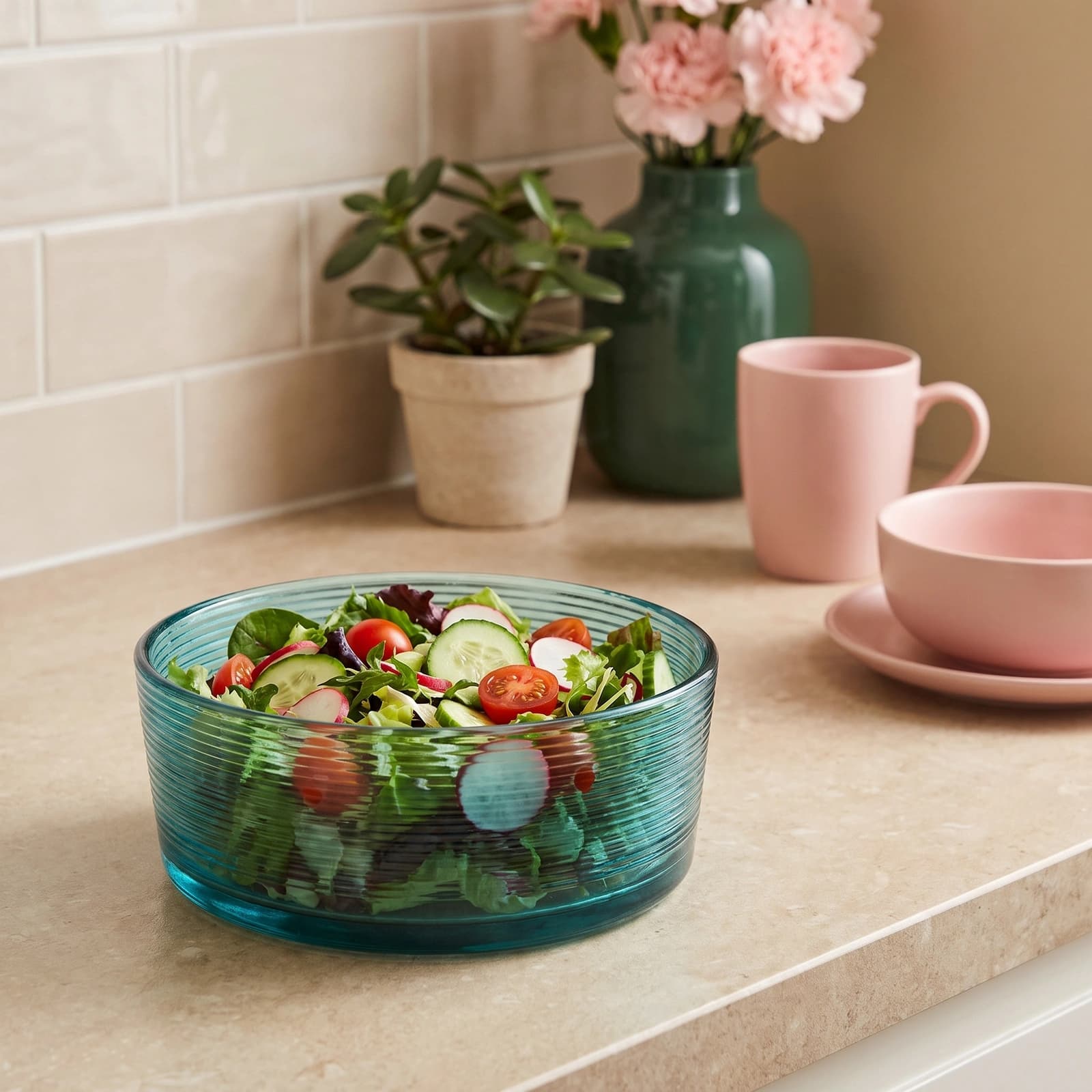 Round teal ribbed glass serving bowl filled with fresh salad on a kitchen counter, with pink mug and vase in background.