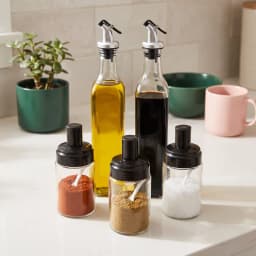 Five clear glass bottles and jars with black and silver tops on a white background. Two tall oil/vinegar bottles, three smal… - view 1