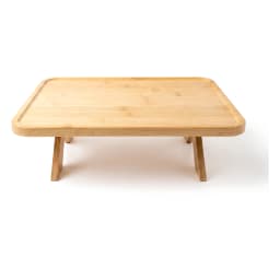 Light brown wood tray with foldable legs on a bedside table, holding a mug, a book, and eucalyptus leaves, next to a bed. - view 2