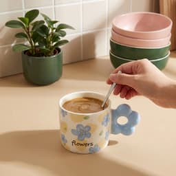 Cream ceramic mug with light blue floral pattern and a unique light blue flower-shaped handle on a white shelf. - view 1