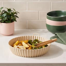 Single round beige silicone air fryer basket liner with wavy bottom and two small handles on a white shelf. - view 1