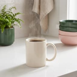 Single plain cream ceramic mug with a handle, sitting on a white shelf against a light background. - view 1