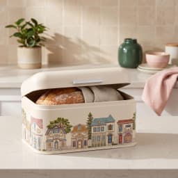 Cream metal bread box with a hinged lid, decorated with a colorful village scene illustration on a white background. - view 5