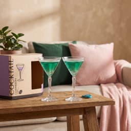 Two clear glass cocktail glasses with green liquid on a wooden table next to a plant and product box, with sofa in backgroun… - view 1