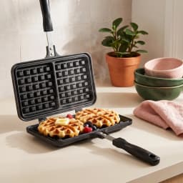 Black granite stovetop waffle maker, open, showing square waffle grids and long black handles on a white background. - view 1