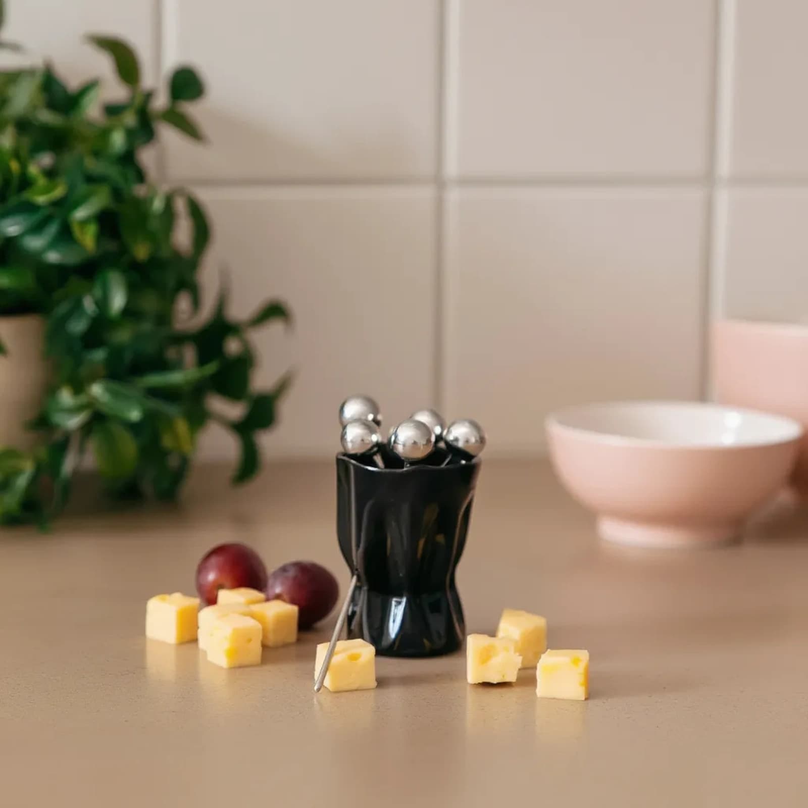 Six silver metal fruit forks with spherical tops in a black ceramic holder, tied with a white ribbon, on a white background.