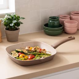 Square ceramic grill pan in beige and cream color, filled with grilled chicken skewers and vegetables on a kitchen counter. - view 1