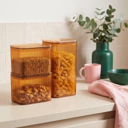 Three amber ribbed acrylic storage containers with square lids, filled with pasta and nuts, on a kitchen counter. - view 1