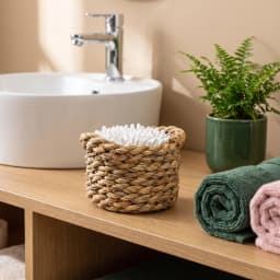 Small round natural khous woven basket holding four lipsticks on a light wooden nightstand next to a bed. - view 1
