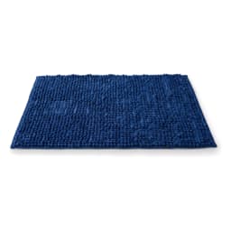 Dark blue rectangular shaggy bath mat on light brown tiled bathroom floor, with a wooden cabinet and rolled towels in backgr… - view 2