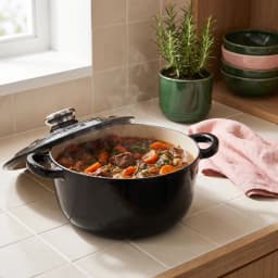 Black cast iron pot with its lid slightly ajar, filled with a steaming beef and carrot stew, on a light tiled kitchen counte… - view 1