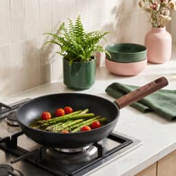 Black granite frying pan with a wooden handle on a gas stove, cooking green asparagus and cherry tomatoes. - view 1