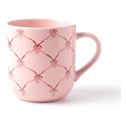 Light pink ceramic mug with a dark pink bow and lattice pattern, filled with a light brown beverage, on a white countertop. - view 2