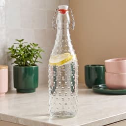 Single clear glass bottle with a textured bumpy surface and a white and orange swing top lid, on a white shelf. - view 1