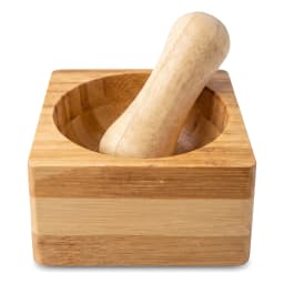 Light brown square wooden mortar and pestle set held in a hand, with kitchen items blurred in the background. - view 2