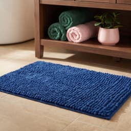 Dark blue rectangular shaggy bath mat on light brown tiled bathroom floor, with a wooden cabinet and rolled towels in backgr… - view 1