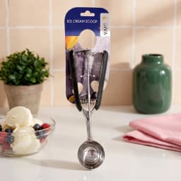 Silver metal ice cream scoop with black rubber handles on a retail card against a light wooden background - view 3