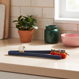 Single dark blue silicone kitchen tongs with a small white tag, lying on a white shelf. - view 1