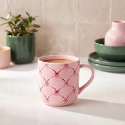 Light pink ceramic mug with a dark pink bow and lattice pattern, filled with a light brown beverage, on a white countertop. - view 1