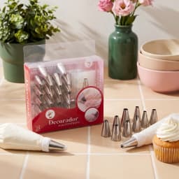 Boxed set of 25 silver metal cake decorating nozzles, a white piping bag, and a coupler, displayed on a shelf. - view 1