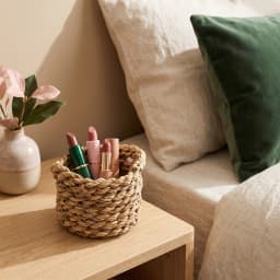 Small round natural khous woven basket holding four lipsticks on a light wooden nightstand next to a bed. - view 3