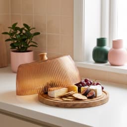 Round wooden base with a ribbed amber acrylic cake dome lid, displaying cheese, crackers, grapes, and figs on a kitchen coun… - view 1