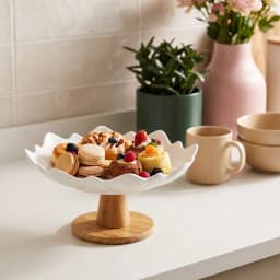 White porcelain serving bowl with a wavy, petal-like rim, sitting on a round light brown wood base, on a white background. - view 4