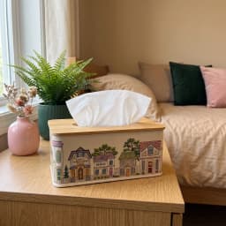 Rectangular cream metal tissue box with a colorful village illustration and a light brown wooden lid, on a white surface. - view 1