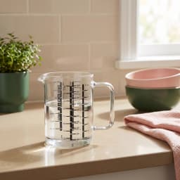 Clear glass measuring jug with a handle and spout, marked with black measurements for litres and fluid ounces, on a white ba… - view 1