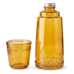 Amber glass bottle with silver cap and matching decorative glass cup on a white shelf against a light background. - view 2