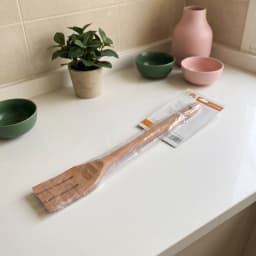 Single brown wooden slotted cooking fork in clear plastic packaging, lying on a white surface in a store shelf. - view 3