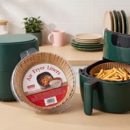Stack of brown round air fryer paper liners in clear packaging next to a dark green air fryer with fries inside. - view 1