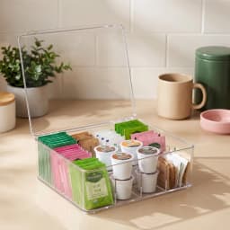 Clear acrylic storage box with a hinged lid and 12 compartments, filled with tea bags and coffee pods on a kitchen counter. - view 1