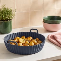 Round dark grey silicone air fryer liner with wavy bottom and fluted sides, featuring two small handles, on a white shelf. - view 1