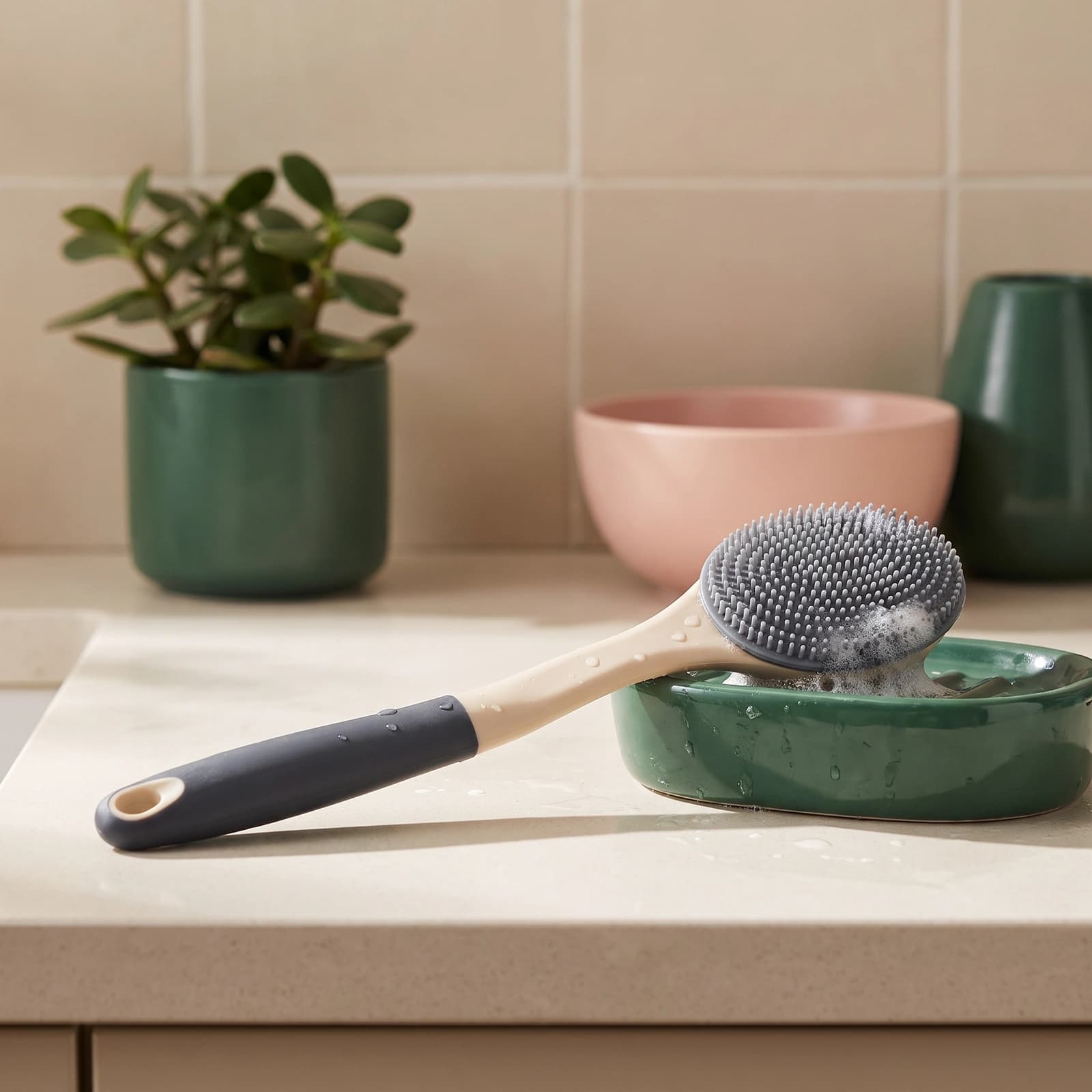 Long-handled silicone bath brush with grey bristles and a beige and dark grey handle, resting on a green soap dish with foam.