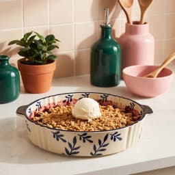 Round cream porcelain baking dish with dark blue leaf patterns and fluted edges, featuring two small handles on a white back… - view 1