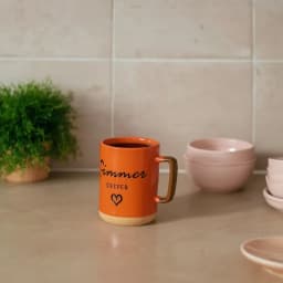 Orange ceramic mug with brown handle and beige base, featuring black 'Simmer Deeper' text and a heart, filled with coffee on… - view 1