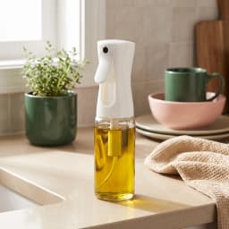 Clear glass oil spray bottle with a white fine mist top, filled with olive oil, standing on a light kitchen counter. - view 1