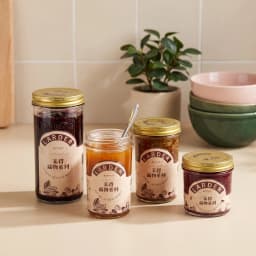 Four clear glass cylindrical storage jars with gold metal lids on a kitchen counter, filled with jam, pasta, and spices. - view 1