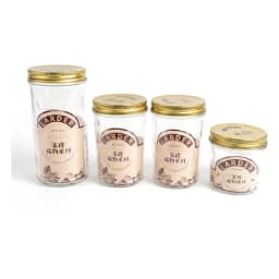 Four clear glass cylindrical storage jars with gold metal lids on a kitchen counter, filled with jam, pasta, and spices. - view 2