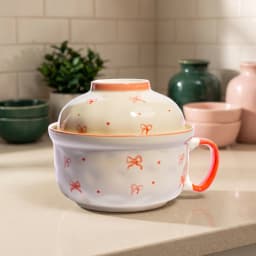 Cream ceramic bowl with a domed lid and handle, decorated with orange bow patterns and dots, on a white background. - view 1