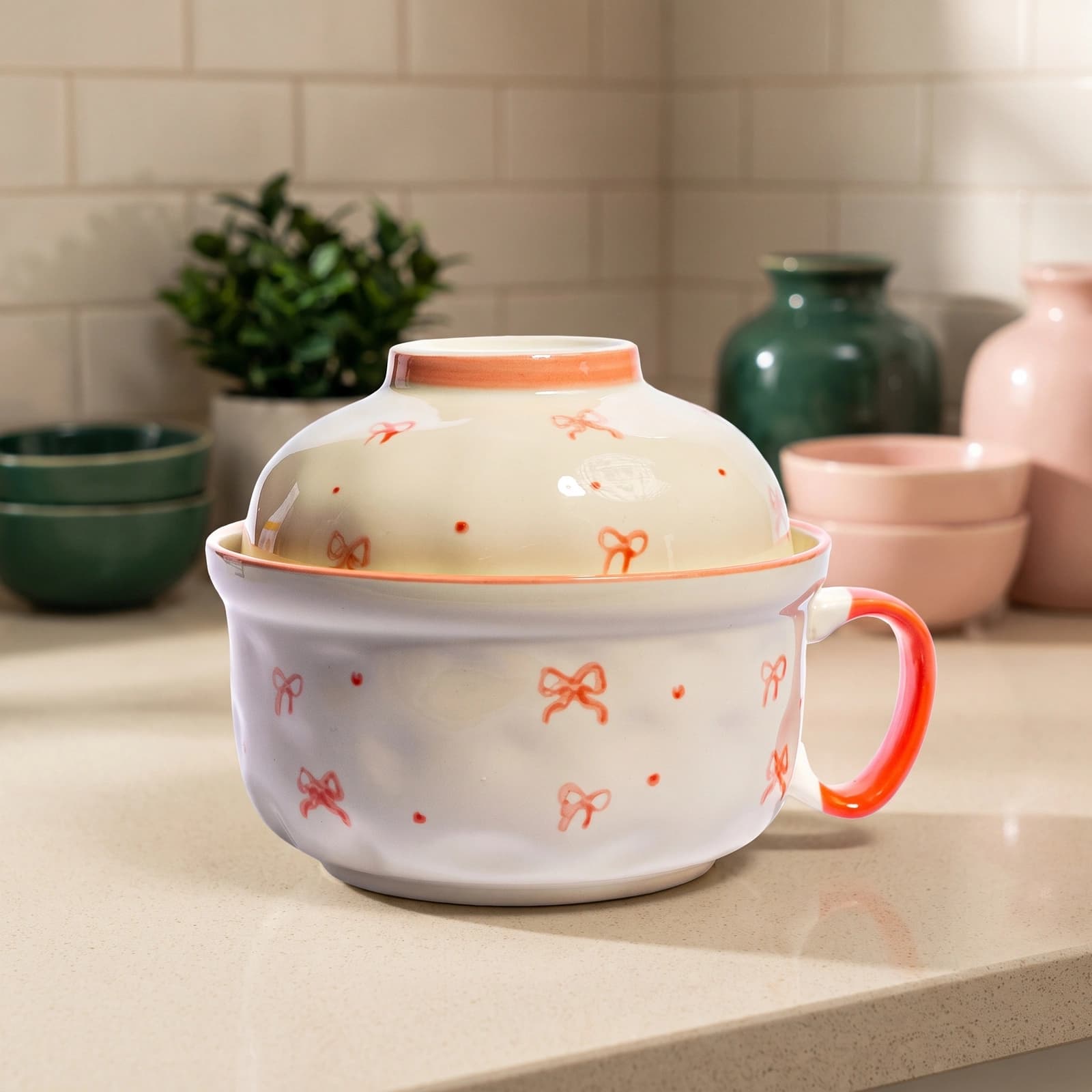 Cream ceramic bowl with a domed lid and handle, decorated with orange bow patterns and dots, on a white background.
