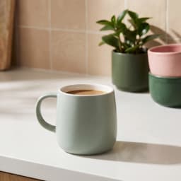 Single light green ceramic mug with a white interior, sitting on a white shelf against a light brown wooden background. - view 1