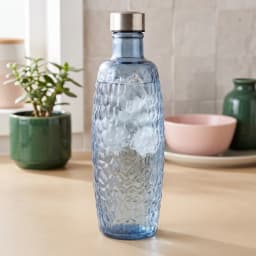 Tall blue glass bottle with a textured honeycomb pattern and a silver metallic cap, standing on a white shelf. - view 1