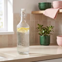 Clear ribbed glass bottle with a white swing top lid, filled with water and lemon slices, on a kitchen counter. - view 1
