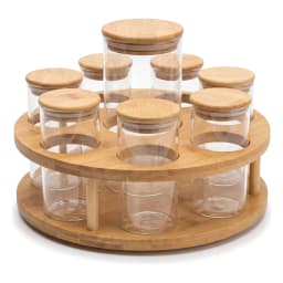 Eight clear glass storage jars with light wood lids on a two-tier rotating wood stand, filled with various spices on a kitch… - view 2