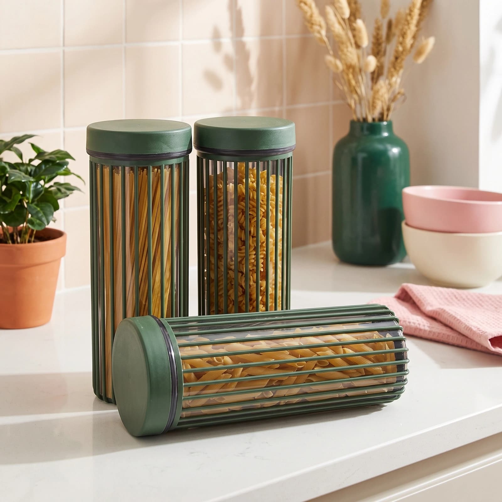Set of three dark green cylindrical acrylic storage jars with clear bodies, filled with pasta, on a white kitchen counter.