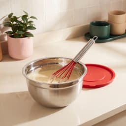 Stainless steel mixing bowl with red whisk and red lid on a light kitchen counter, next to a pink potted plant. - view 8